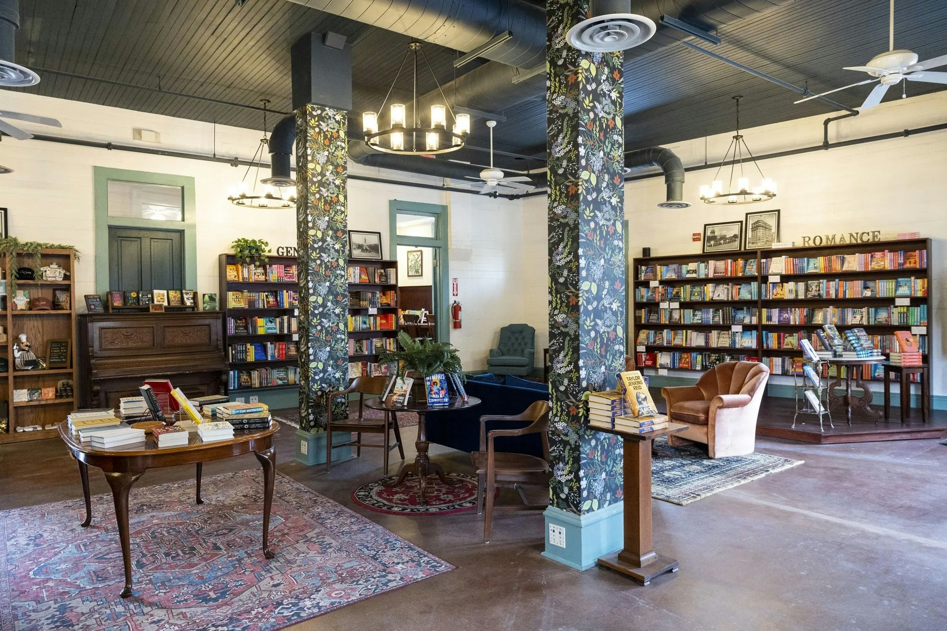 Interior of a cozy independent bookstore with wooden bookshelves filled with colorful books, vintage-style furniture, floral-patterned columns, and warm lighting creating an inviting atmosphere.