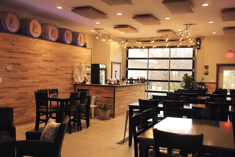 A warm and inviting wine tasting room featuring wooden decor, hanging string lights, wine barrels on display, and a bar with seating. Large windows let in natural light, creating a cozy ambiance.