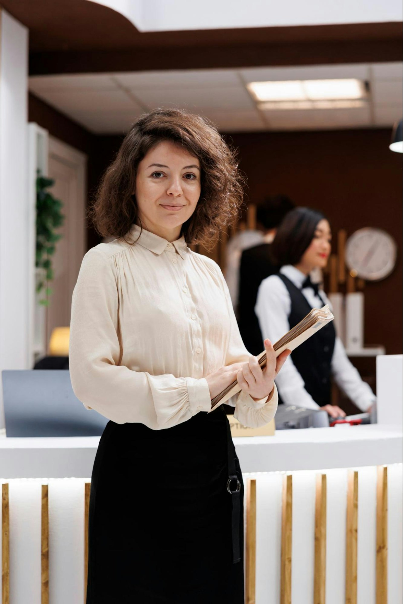 A confident woman with curly brown hair, wearing a cream blouse and black skirt, stands at a hotel reception desk holding a folder. Behind her, two hotel staff members in formal uniforms assist guests. The reception area has a modern design with warm lighting and a professional atmosphere.