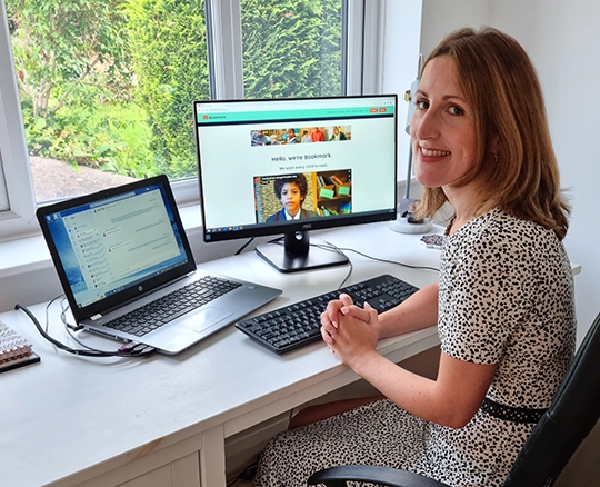 Katie sits at her desk with her laptop and monitor, looking at Bookmark's website
