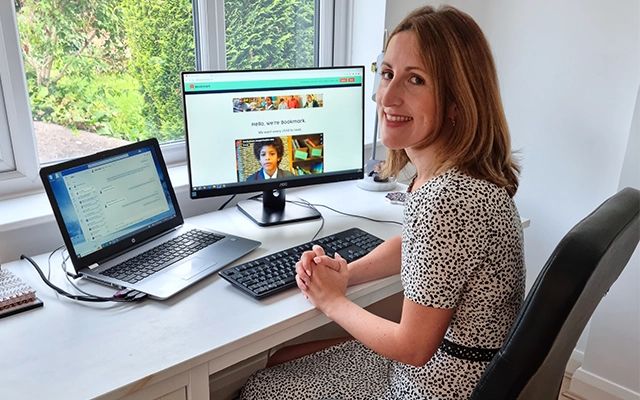 Katie sits at her desk with her laptop and monitor, looking at Bookmark's website