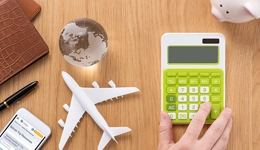 A birds eye view of a desk, with a person's hand hovering over a calculator and a piggy bank, globe, toy plane, mobile phone, pen and diary surrounding