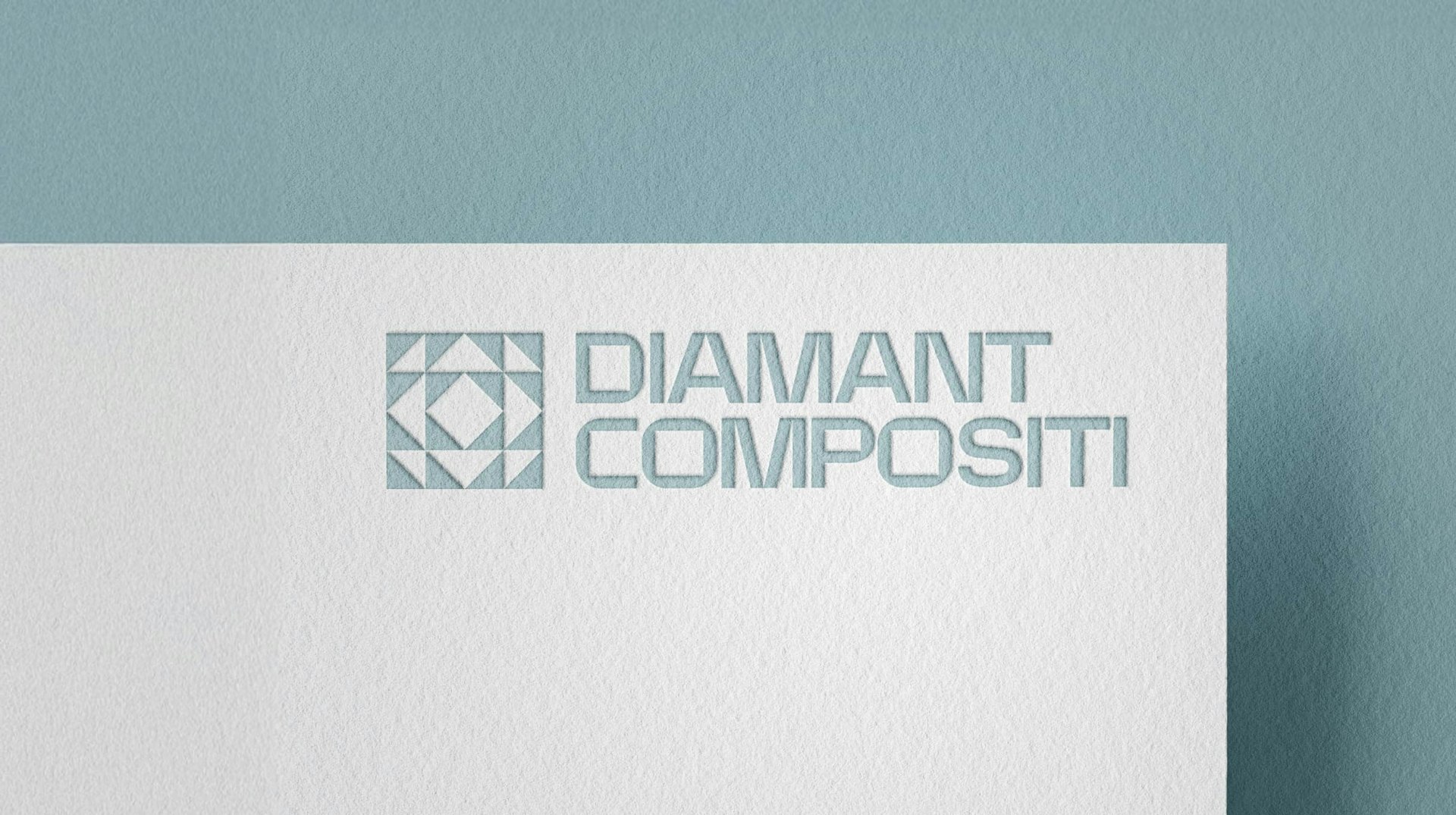 L'immagine mostra un logo per Diamant Compositi in lettere blu-grigie su una superficie bianca testurizzata. Il logo include un motivo geometrico a forma di diamante a sinistra del testo. Lo sfondo è di un blu tenue e smorzato.