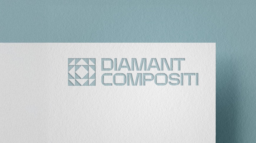 L'immagine mostra un logo per Diamant Compositi in lettere blu-grigie su una superficie bianca testurizzata. Il logo include un motivo geometrico a forma di diamante a sinistra del testo. Lo sfondo è di un blu tenue e smorzato.