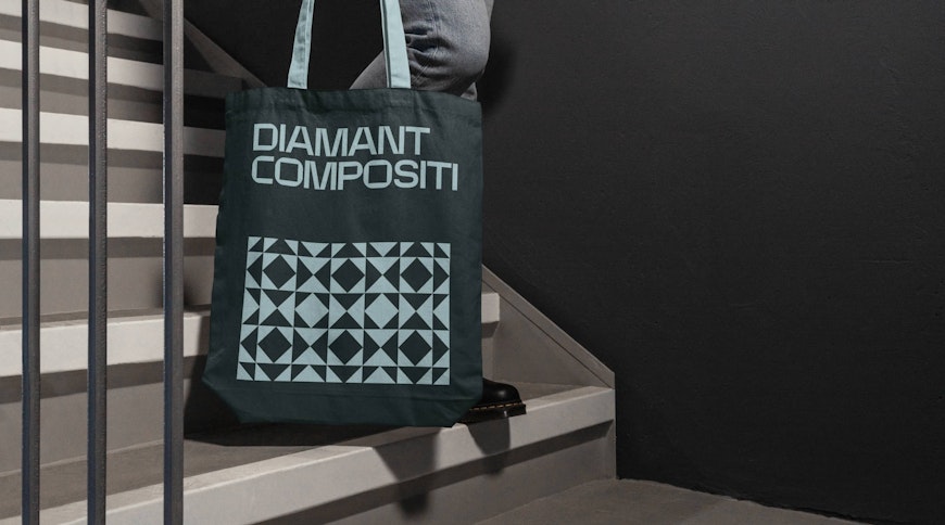 Persona che scende le scale, con in mano una tote bag verde scuro con DIAMANT COMPOSITI e un motivo geometrico. Lo sfondo è un muro grigio scuro e le scale sono bianche con ringhiere nere.