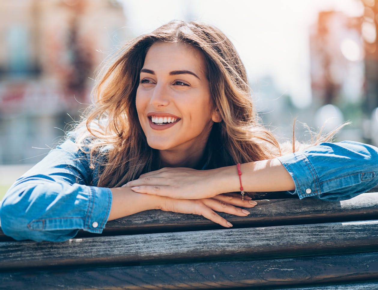 Woman leaning on wooden fence with city blurred out in background.