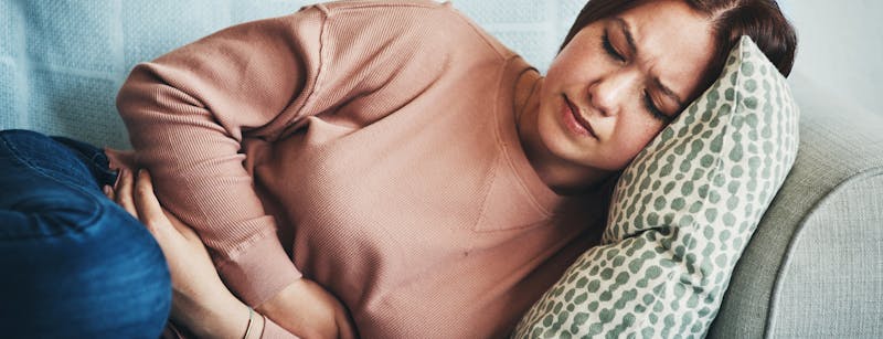 Woman with a look of distress, sits on a couch holding her stomach.