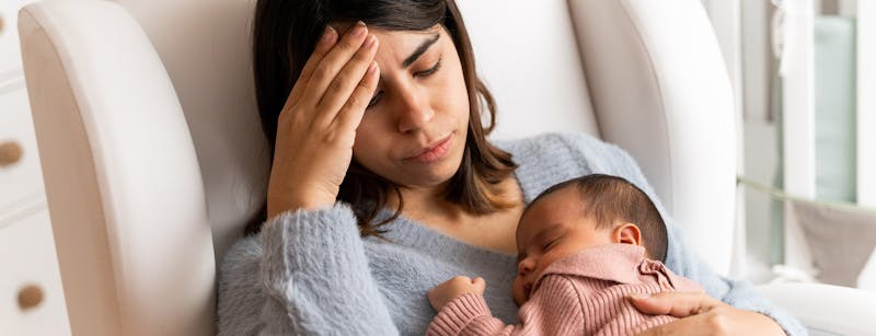Woman in a chair holding her baby in one arm and her head with her other hand.