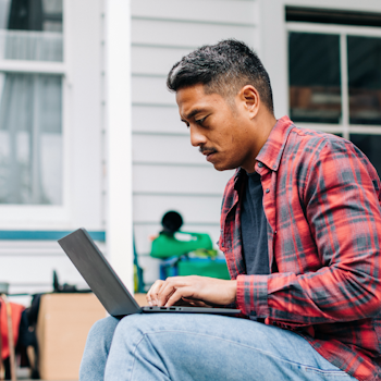 Young man sitting outside weatherboard house on laptop.