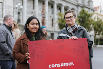 Two women holding a red Consumer sign outside Parliament.
