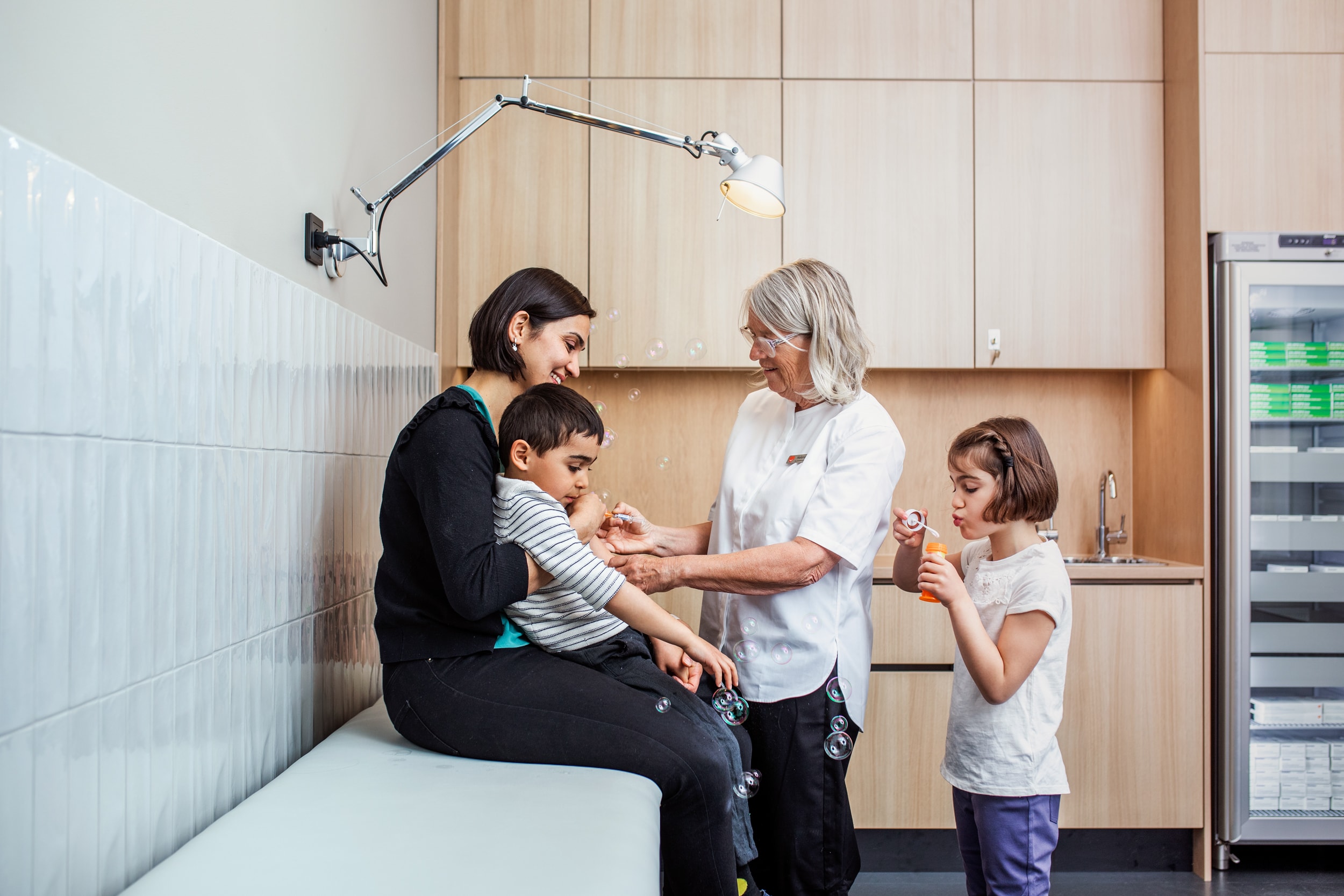 Sjuksköterska ger vacccinspruta samtidit som mamma håller honom och dotter blåser såpbubblor