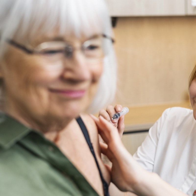 Äldre kvinna till vänster får en spruta i överarmen av sjuksköterska som sitter till höger.