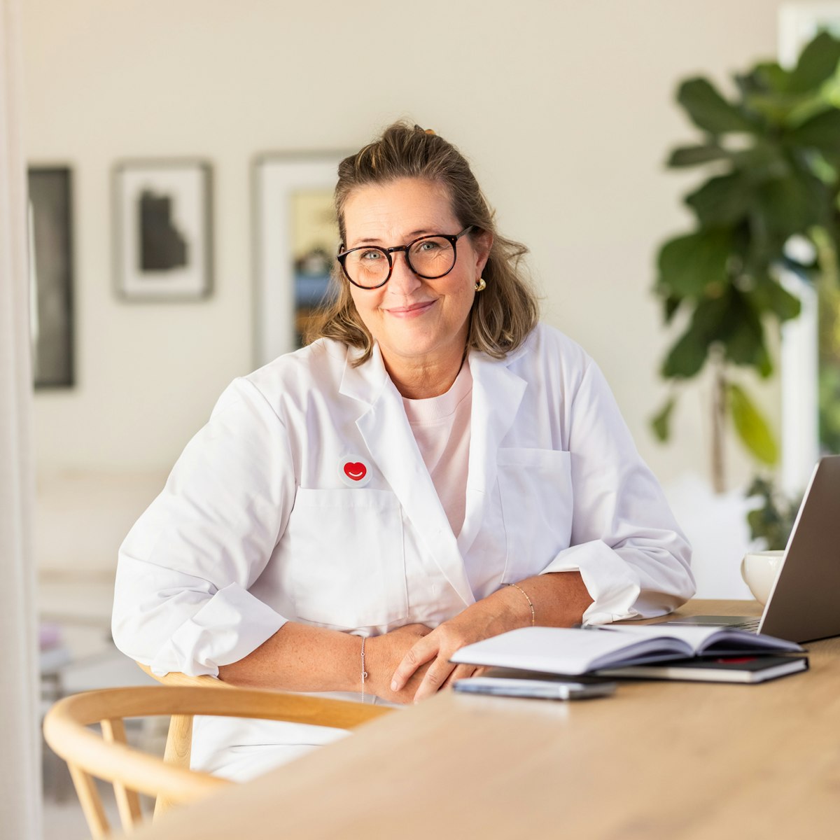 Kvinnlig läkare sitter med rock framför en dator hemma och kollar leendes in i kameran. Hon har glasögon på sig. Bakom datorn står en vattenflaska med Min Doktor logga och vid sidan av datorn ligger ett anteckningsblock.