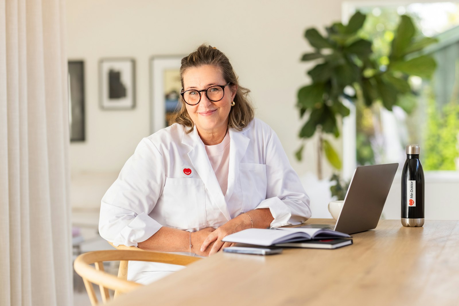 Kvinnlig läkare sitter med rock framför en dator hemma och kollar leendes in i kameran. Hon har glasögon på sig. Bakom datorn står en vattenflaska med Min Doktor logga och vid sidan av datorn ligger ett anteckningsblock.