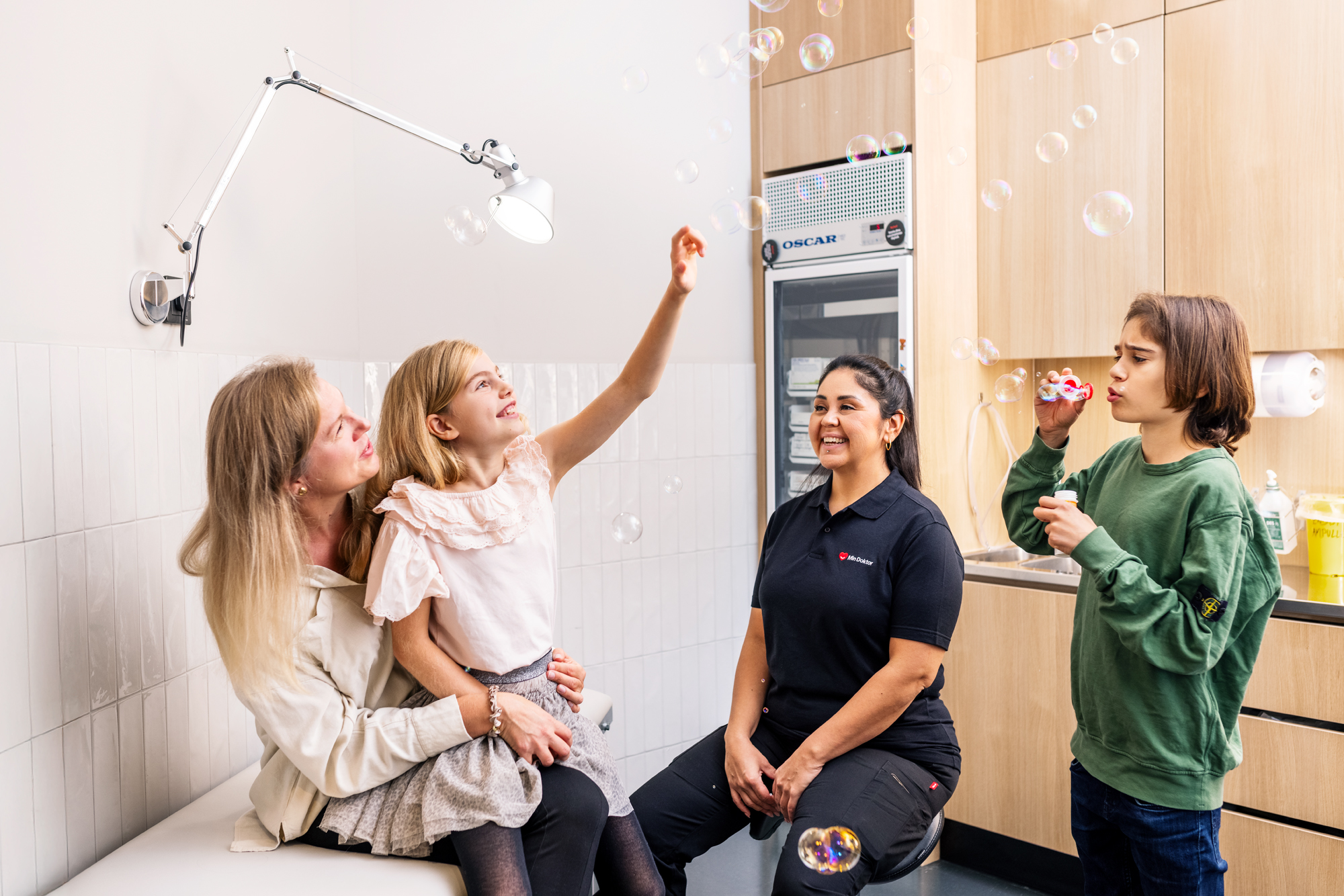 Kvinna till vänster sitter på en brits med en flicka i knät som tittar på såpbubblor. En sjuksköterska sitter till höger på en stol och kollar leendes på och en pojke står och blåser bubblor till höger om henne.