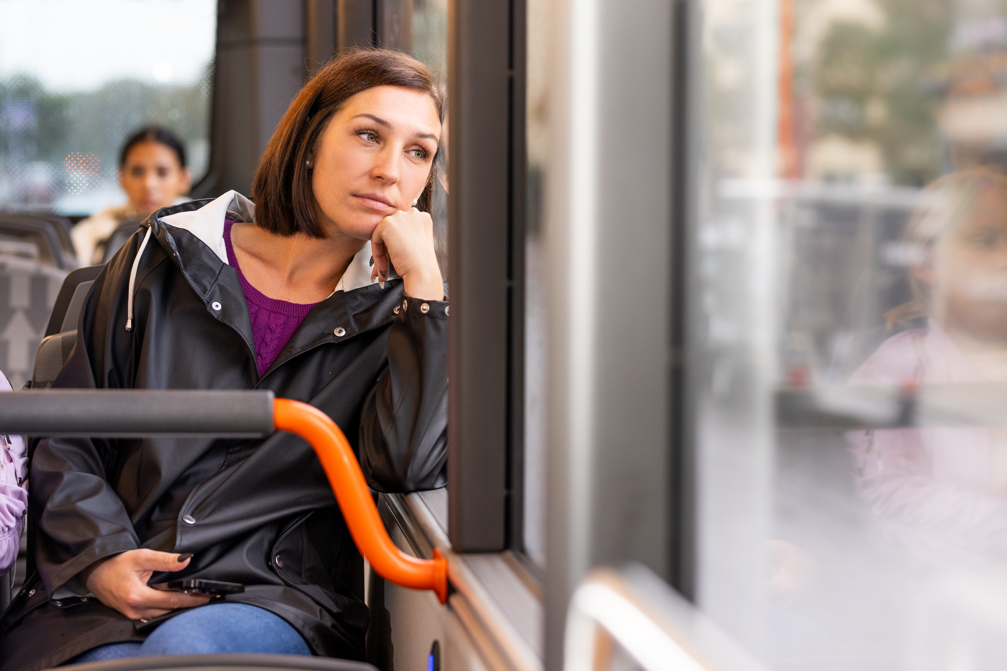 Kvinna sitter på en buss och kollar ut ur fönstret till höger. Har en mobil i vänster hand.