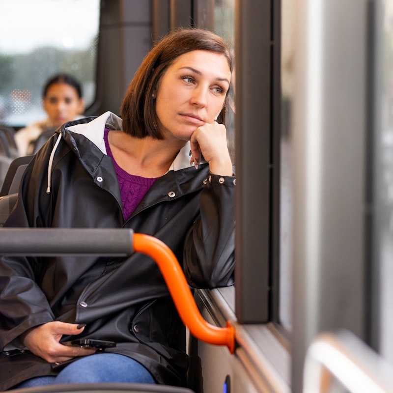 Kvinna sitter på en buss och kollar ut ur fönstret till höger. Har en mobil i vänster hand.