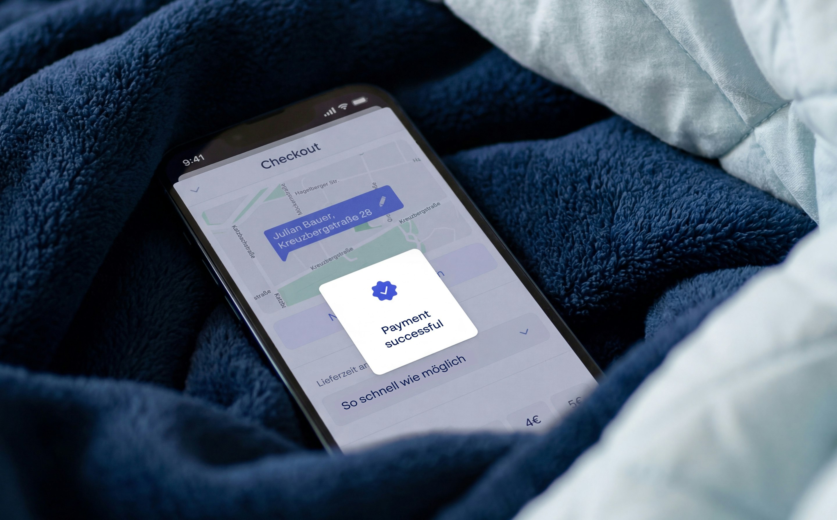 Puls app checkout screen shown on a smartphone resting on a navy fleece blanket, with a 'Payment successful' confirmation card and German delivery details.