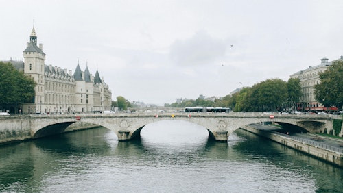 Malerischer Blick auf eine historische Brücke über den ruhigen Fluss mit klassischer Pariser Architektur im Hintergrund.