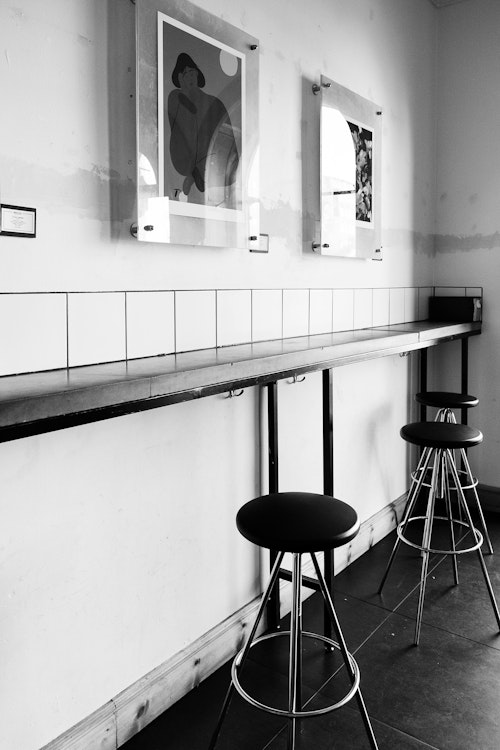 Schwarz-Weiß-Foto eines leeren Café-Interieurs mit Hockern und Wandkunst.