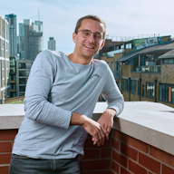 Headshot of a man in a grey shirt resting on a brick wall balcony overlooking skyscrapers