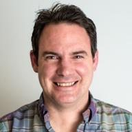 A person with short dark hair wearing a plaid shirt is smiling at the camera against a plain background.