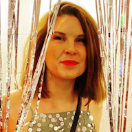 A person with shoulder-length hair and red lipstick stands among metallic streamers, wearing a sequined top.