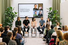 A panel of five speakers seated on high stools in front of an audience at an event titled “Breaking ground: bringing AI-powered products to market in large organisations.” One speaker is holding a microphone while speaking. A large screen behind them displays the event title and speaker names, with logos of OpenAI, Planes, and Techspace. The room is bright with large plants on either side and attendees listening attentively.