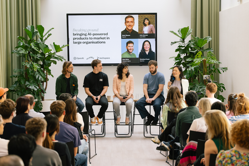 A panel of five speakers seated on high stools in front of an audience at an event titled “Breaking ground: bringing AI-powered products to market in large organisations.” One speaker is holding a microphone while speaking. A large screen behind them displays the event title and speaker names, with logos of OpenAI, Planes, and Techspace. The room is bright with large plants on either side and attendees listening attentively.