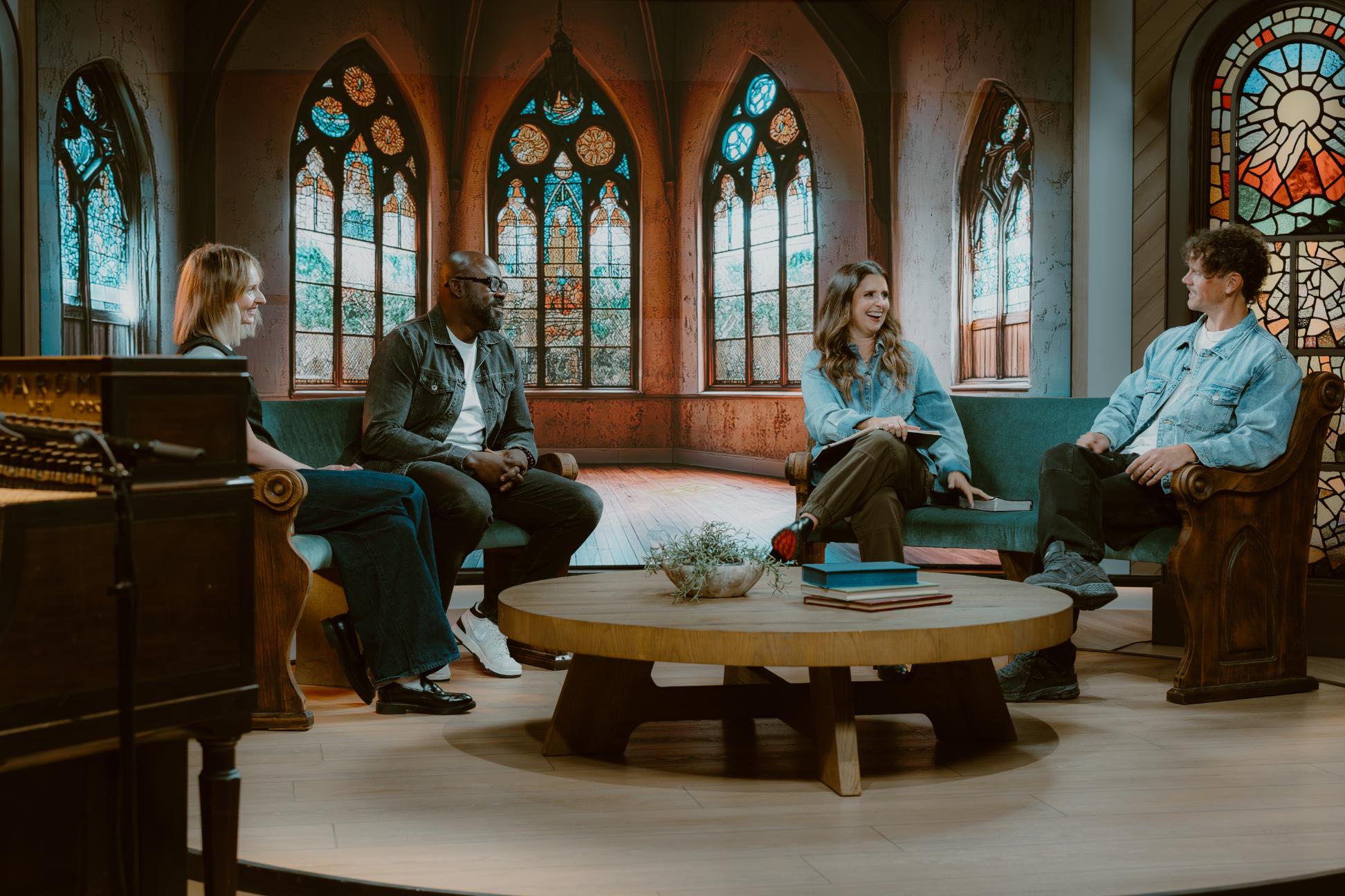 Four adults sit together in a studio styled like a church, talking and smiling around a round coffee table.