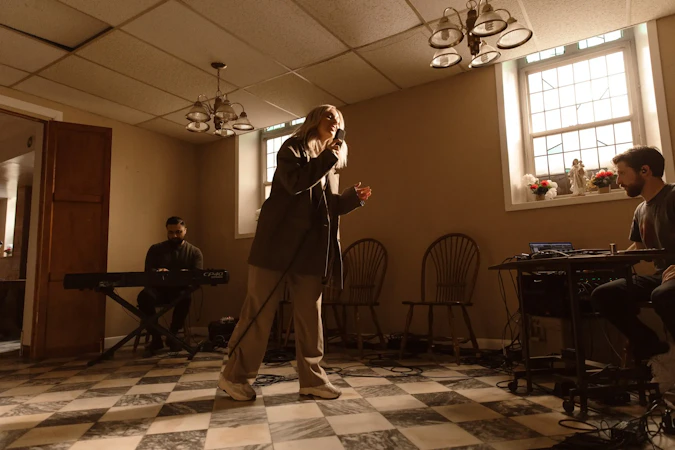 Tiffany standing in the middle of a room, singing into a microphone. There is a man sitting behind her playing a piano and a sound engineer to her right.