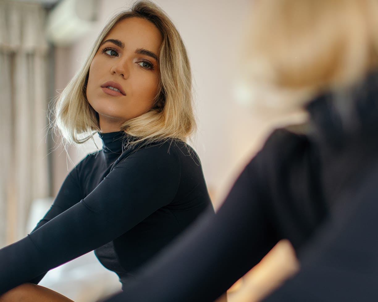 blonde woman looking into a mirror wearing a black turtle neck