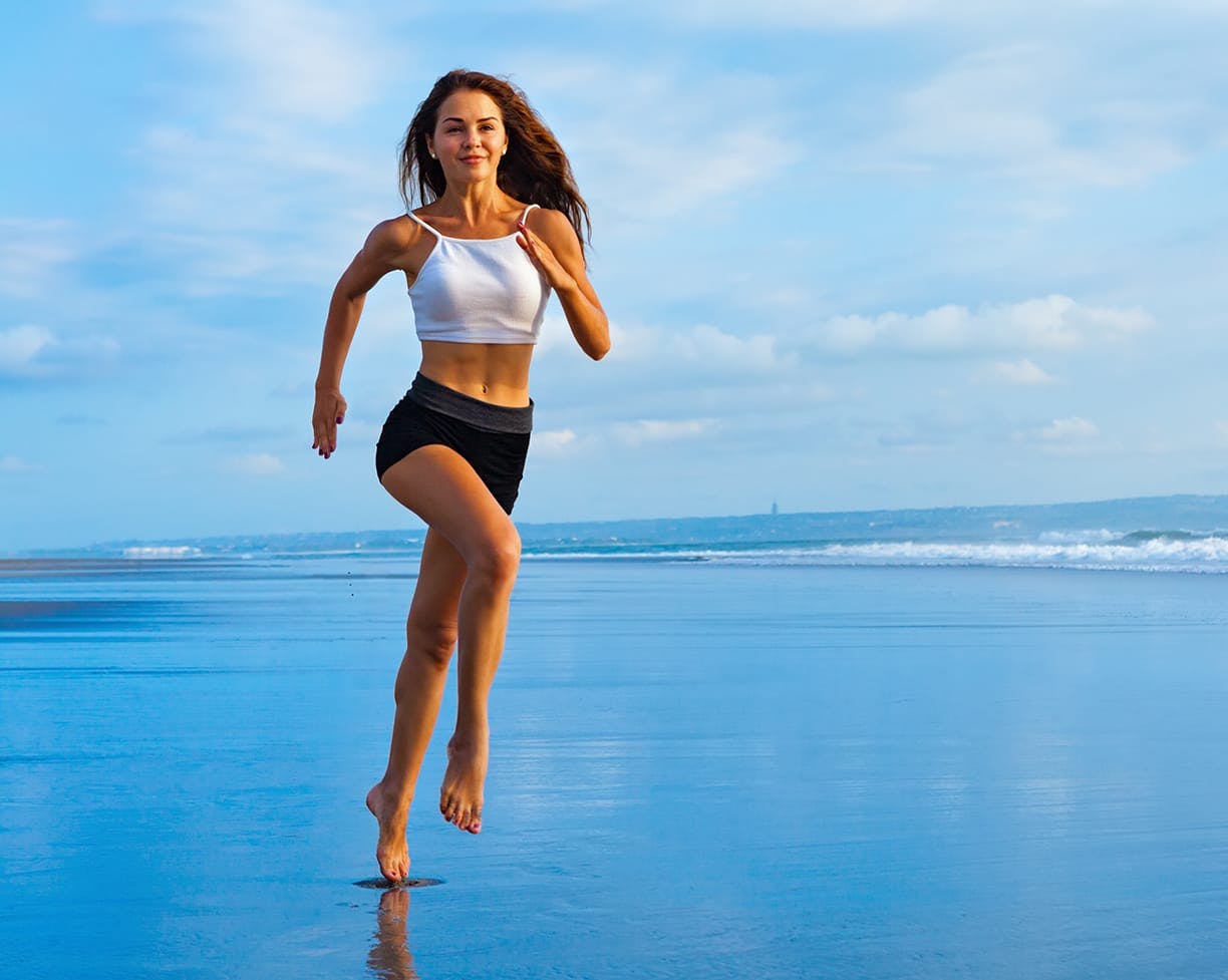 a brunette woman running on the beach in a white tank top and black shorts