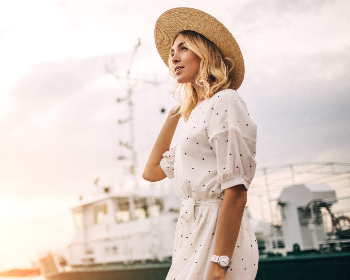 a blonde model in a white dress and sun hat standing by a boat