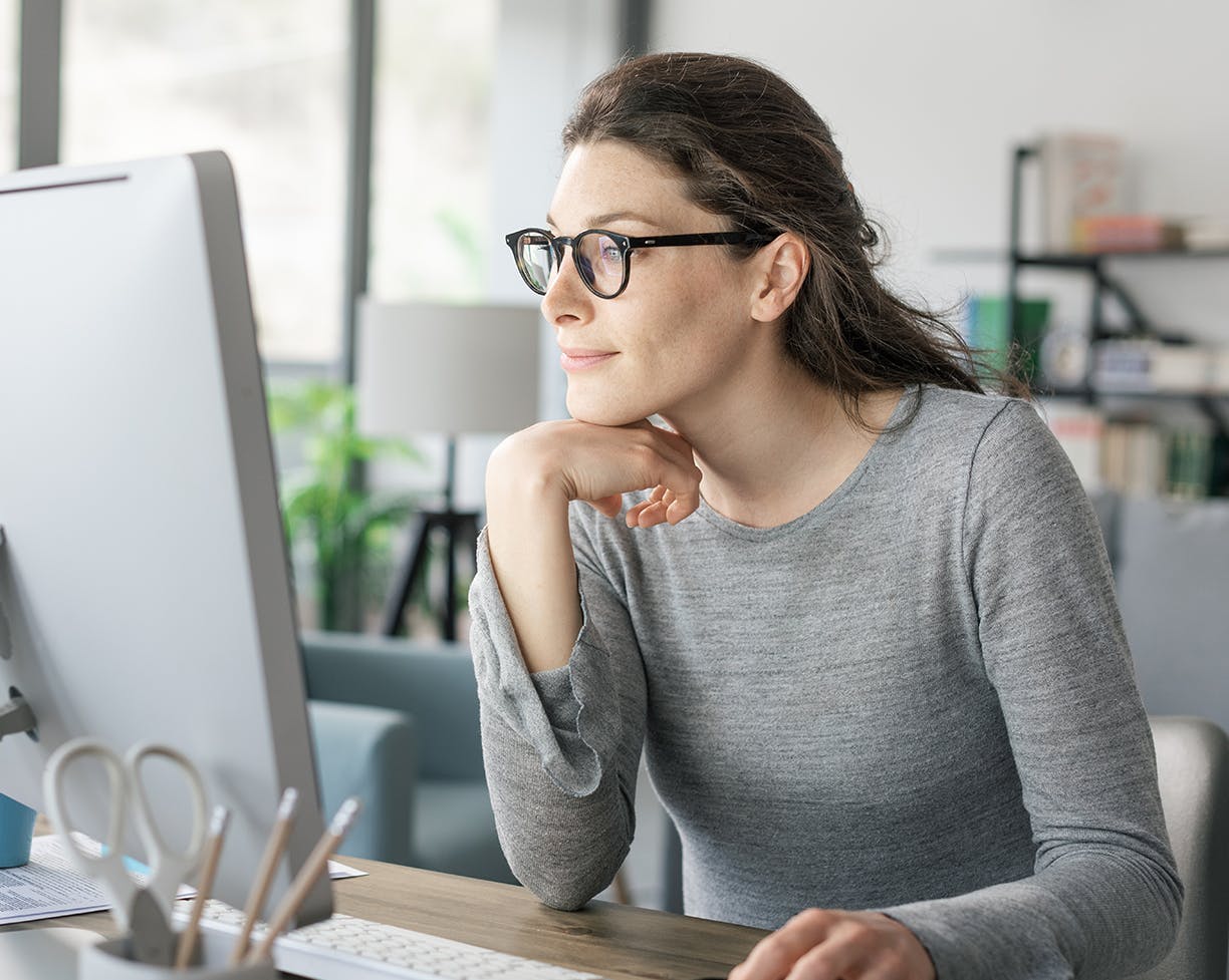 a brunette woman in glasses in a grey long sleeve looking at a computer screen