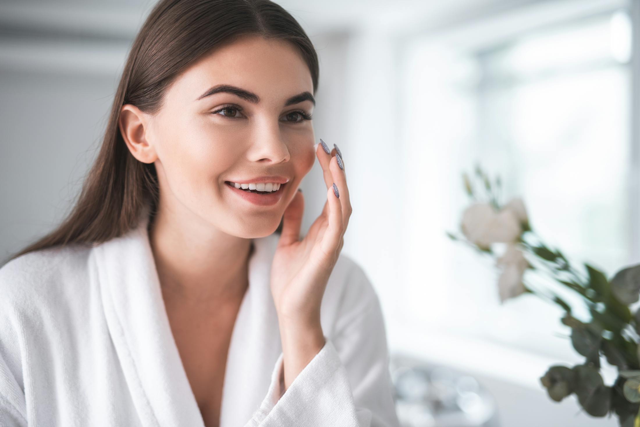 a brunette woman looking into the mirror smiling and touching her cheek