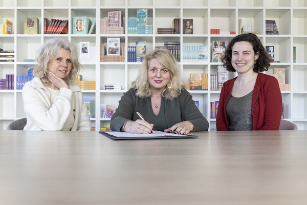 3 vrouwen aan een tafel, middelste vrouw ondertekent een papier