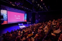 Gevulde theaterzaal gericht op het podium met een groot scherm, daarop staat het logo van de Mediafederatie