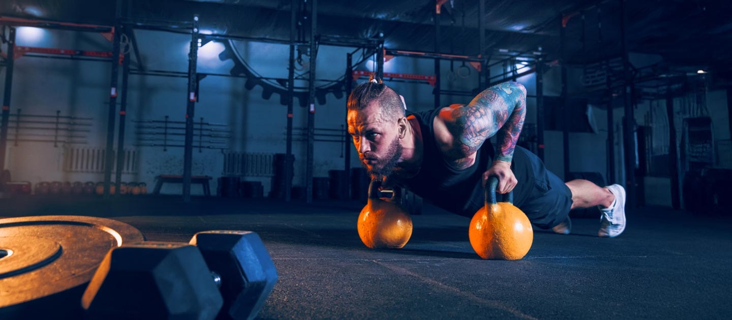 Athletic man performing pushups on two yellow kettlebells in a dark gym environment.