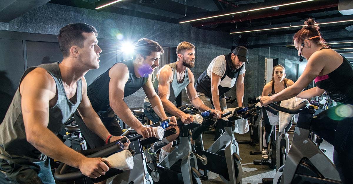 Group of participants riding stationary bikes during an indoor cycling spin class led by a fitness instructor.