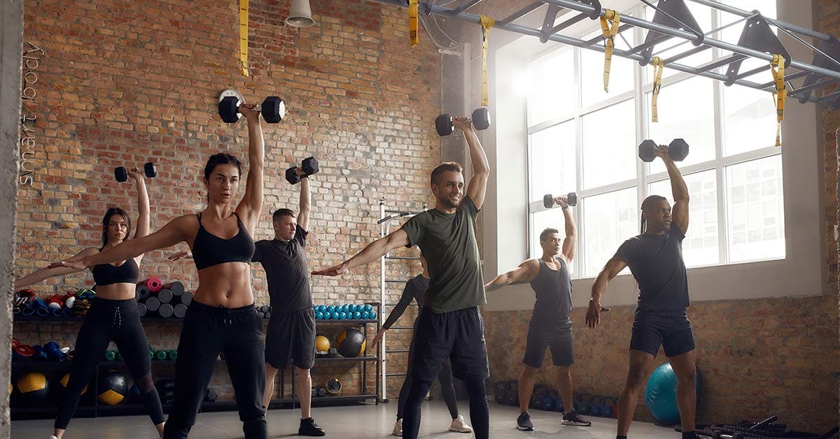 Group of participants performing single-arm overhead dumbbell presses during a group fitness class in a gym studio.