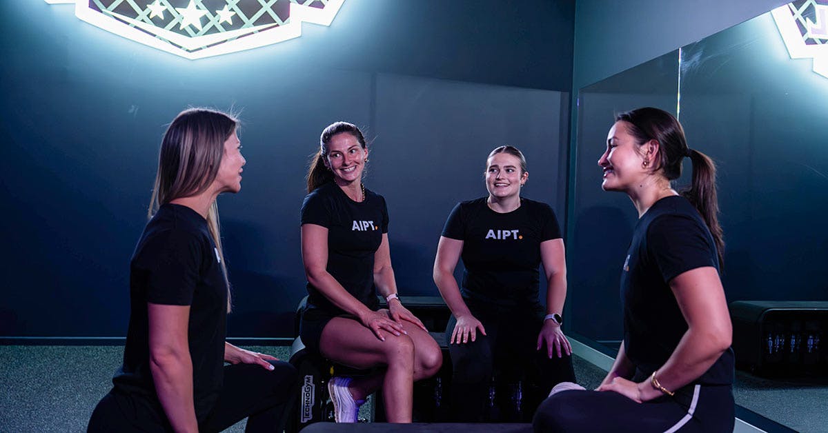 Four female AIPT fitness students and mentors wearing branded black shirts talking together in a gym studio setting.