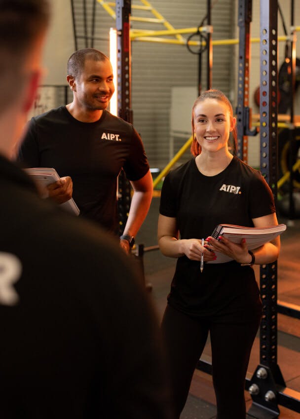 Male and female AIPT fitness students holding training notebooks and smiling during a gym mentoring session.