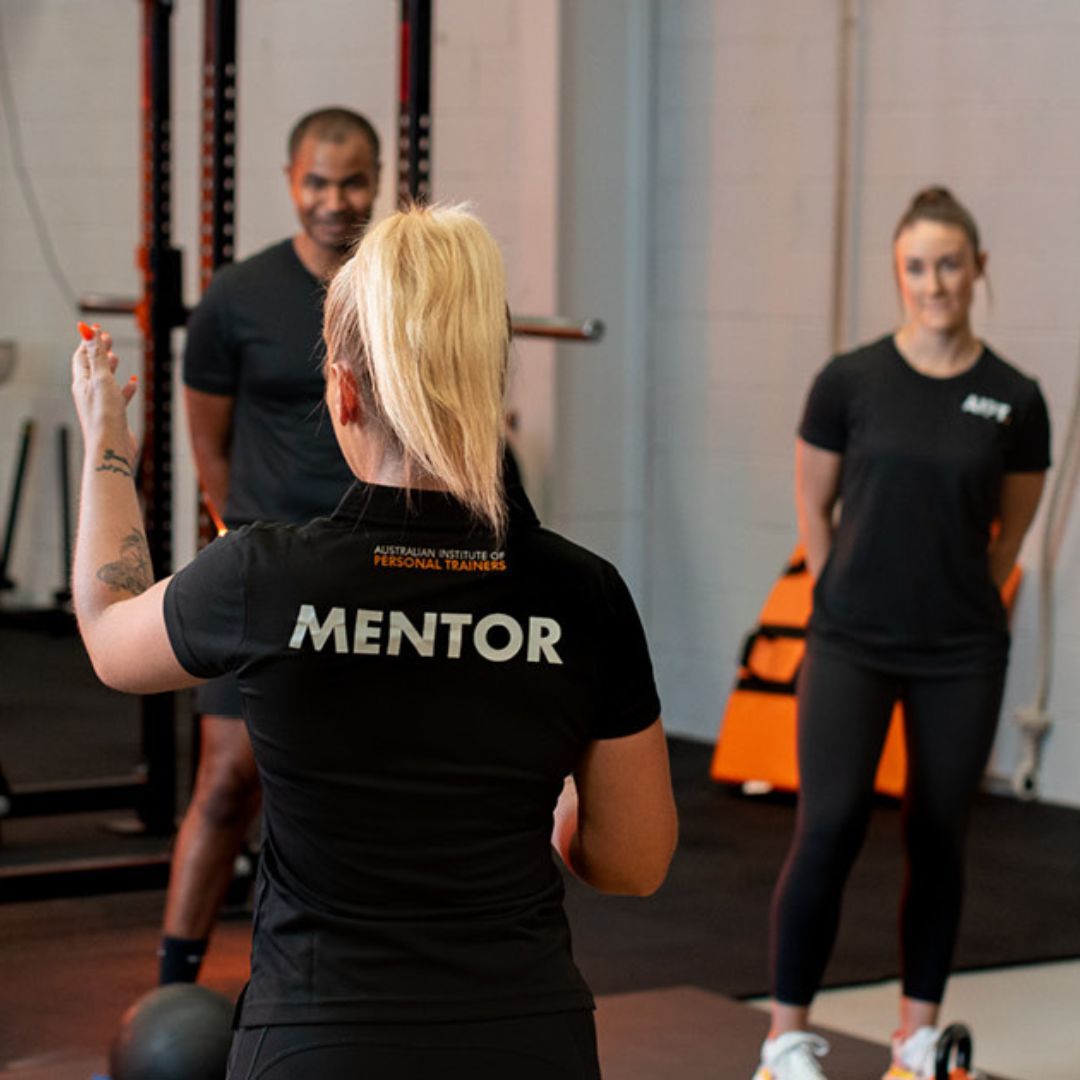 AIPT mentor wearing a labeled shirt instructing two fitness students during practical gym training.