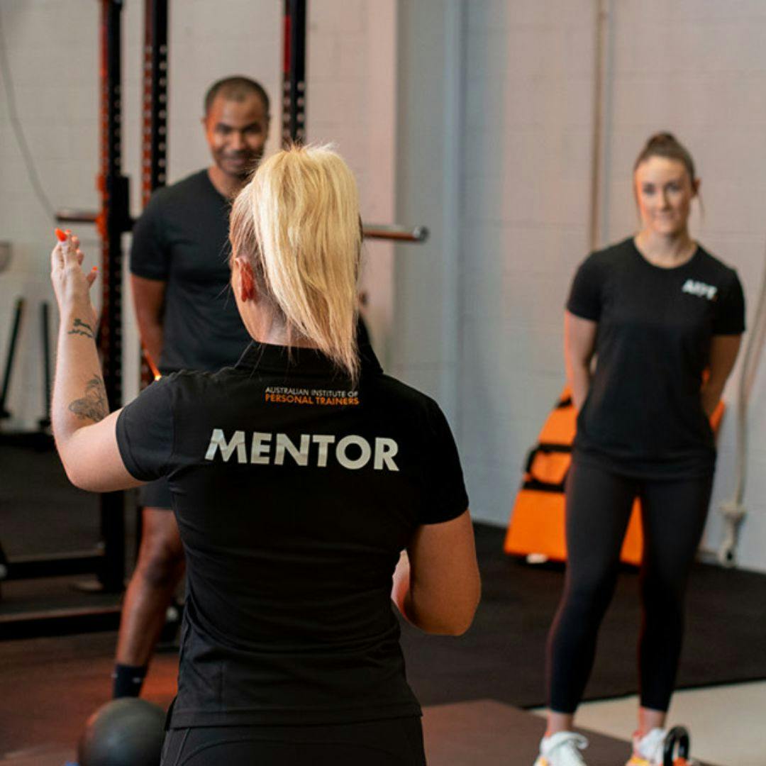 AIPT mentor wearing a labeled shirt instructing two fitness students during practical gym training.