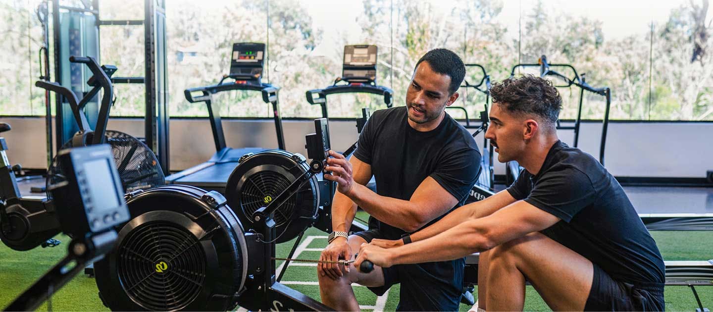 Male AIPT mentor instructing a student on rowing machine technique during practical gym training.