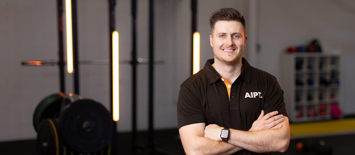 Smiling male AIPT fitness educator wearing a branded black shirt standing with arms crossed in a gym studio.