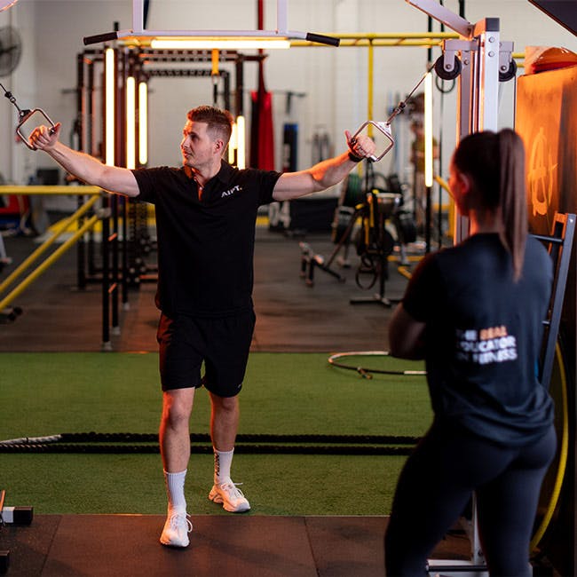 Male AIPT trainer demonstrating a cable fly exercise to a female student in a gym studio.