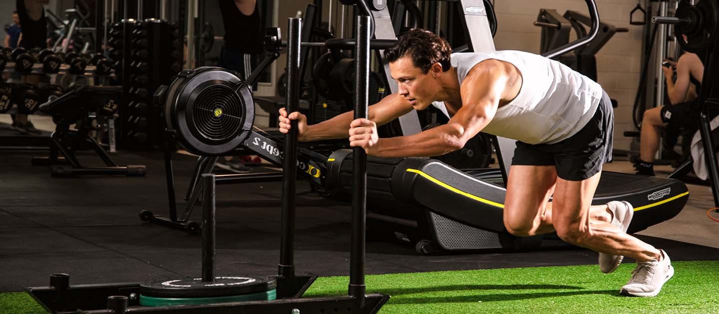 Male athlete pushing a heavy weighted sled across indoor gym turf during a fitness training session.