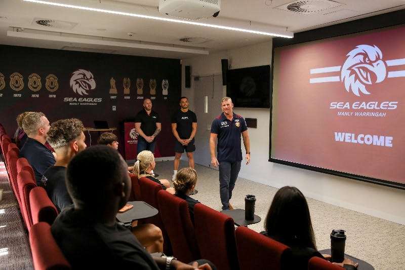 A presenter speaks to AIPT fitness students seated in an auditorium at the Manly Warringah Sea Eagles facility during a practical masterclass.
